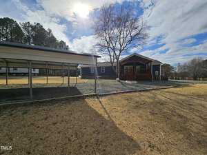 A wooden house with a covered porch is situated in a yard with dry grass. A metal carport stands to the left. The sky is partially cloudy, and the sun is visible, creating shadows across the scene.