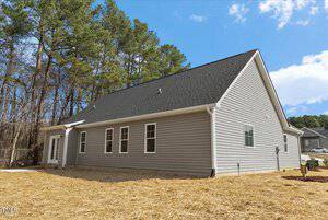 Single-story gray house with a sloped black roof, several windows, and a white back door. The yard is covered in straw, bordered by trees on the left and a partly cloudy blue sky above.