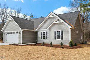 A modern single-story gray house with white trim, featuring a two-car garage, dark shutters, and decorative gables. The yard is recently landscaped with small shrubs and straw mulch, surrounded by trees under a cloudy blue sky.