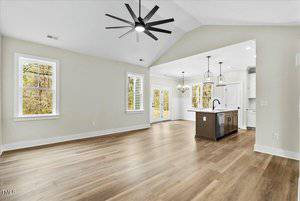 Spacious open-concept living area with light wood floors, featuring a large ceiling fan and recessed lighting. The room flows into a modern kitchen with pendant lights, white cabinets, and a central island. Two sets of windows provide natural light.