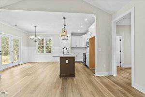 A modern kitchen with white cabinets, stainless steel appliances, and a central island. Light wood flooring extends into a dining area with large windows and glass double doors. Two pendant lights hang over the island. Walls are painted light gray.