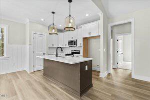 Modern kitchen with a central island featuring a dark base and a white countertop. Two pendant lights hang above. White cabinets and stainless steel appliances are in the background. Light wood flooring and neutral walls complete the look.