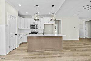 Modern kitchen with a central island featuring a light wood base and white countertop. Stainless steel appliances, including a microwave and stove, are installed in white cabinetry. Two pendant lights hang above the island. Wood flooring is visible.
