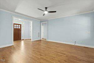 A vacant room with light blue walls, white trim, and hardwood flooring. There is a dark wooden door with glass panels next to the entryway, and a black ceiling fan. An open doorway leads to another room. Electrical outlets are visible on the wall.