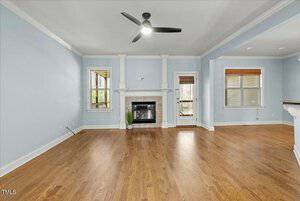 A spacious living room with light blue walls, hardwood floors, and a ceiling fan. The room features a white framed fireplace, two windows flanking the fireplace, and a glass door leading outside. A partial view of a bar counter is on the right.