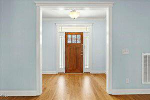 A hallway with light blue walls leads to a wooden front door with glass panels. The door is framed by narrow vertical windows on either side. Above the door hangs a ceiling light fixture. The floor is hardwood.