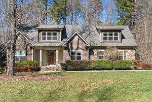 Located in Franklinton, this two-story house at 3659 Brooks Bend features gray shingles, brown siding, and a striking stone accent wall. A welcoming porch with twin columns and double doors opens to a row of bushes, framed by lush trees and a grassy lawn in the background.