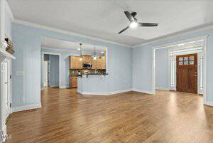 A spacious, empty living room with light blue walls and wooden flooring. It features a ceiling fan, a view into a kitchen with wooden cabinets and a breakfast bar, and a wooden front door with glass panels on the right.