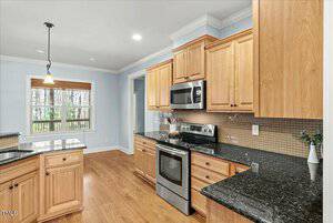 A kitchen with wooden cabinets, black granite countertops, and a stainless steel oven and microwave. A window with blinds is on the left wall. A vase with greens is to the right of the stove. The floor appears to be hardwood.