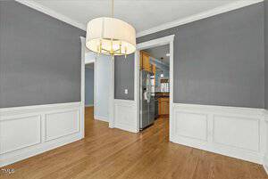 A dining area with wooden flooring, gray walls, and white wainscoting. A large, round light fixture hangs from the ceiling. The room opens into a kitchen with stainless steel appliances visible.