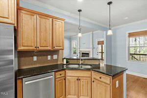 Kitchen interior with wooden cabinets and a black granite countertop. A stainless steel dishwasher is below the counter. Pendant lights hang above, and a window reveals a view of trees through white-trimmed framing.