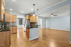 Spacious kitchen and living area with wooden floors and light blue walls. The kitchen features wooden cabinets, a tiled backsplash, and a central island. A ceiling fan is visible in the living area. Recessed and pendant lighting is seen throughout.