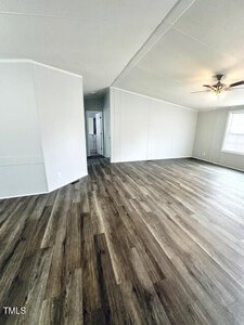 An empty room with light gray walls and dark wood laminate flooring. A ceiling fan with a light is on the right. A hallway leads to another part of the house. A window is on the right wall, letting in natural light.