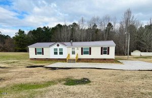 A manufactured home with a white exterior and red trim, set on a grassy field. The front has a small porch with yellow railings and a gravel driveway. Leafless trees are in the background under a cloudy sky.