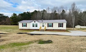 A one-story modular home with light-colored siding and a metal roof is situated on a grassy plot. It has a small wooden porch with steps leading to the front entrance. Trees and a gravel driveway are visible in the background.