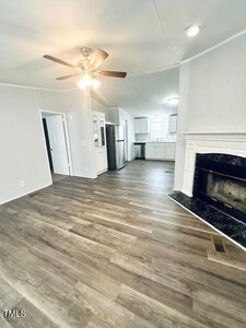 A modern living area with wood flooring, white walls, and a ceiling fan. A fireplace is on the right, and a kitchen with stainless steel appliances is visible in the background. There are doors leading to adjacent rooms.