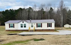 A single-story, beige manufactured home with a brown metal roof and red shutters sits on a grassy lot. There is a gravel driveway and steps leading to the front door. Trees and a small shed are in the background under a partly cloudy sky.