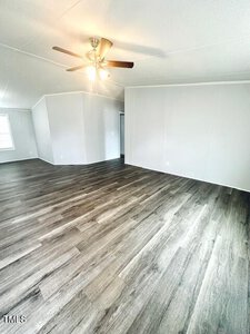 A spacious room with light gray walls and dark wood flooring. The ceiling features a fan with a light. The room has one visible wall outlet and a doorway leading to another room. Natural light enters from a window in the corner.