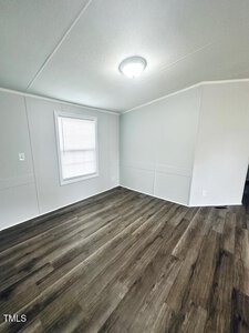 A small, empty room with white walls and a ceiling light. There is a window with closed blinds on one wall. The floor is covered with dark wood-style laminate. The room appears to have a slightly sloped ceiling.