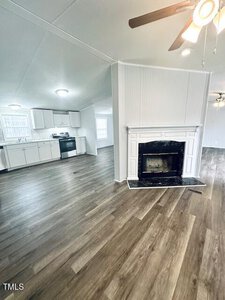 A spacious interior features an open living area with wood-look flooring. There is a white fireplace with a black hearth in the foreground. The kitchen in the background has white cabinets and stainless steel appliances. Ceiling lights and a fan are visible.