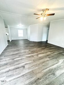A spacious, empty room with light gray walls and dark wood flooring. A ceiling fan with lights is mounted above. There are several open doorways leading to adjacent spaces, and a window in the background.