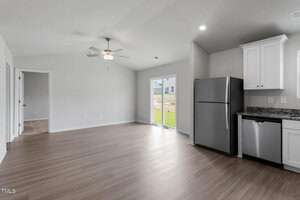 A modern, unfurnished kitchen and living area with light wood flooring. It includes white cabinets, a stainless steel refrigerator, and dishwasher. A ceiling fan is visible above, and there is a large windowed door leading outside.