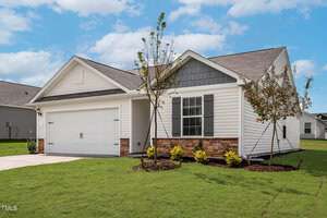 This charming single-story house at 103 Flue Court in Oxford features a white exterior, gray shingles, and stone accents. With a two-car garage, black shutters, and a cozy porch, the landscaped front yard boasts young trees and shrubs under a partly cloudy sky.