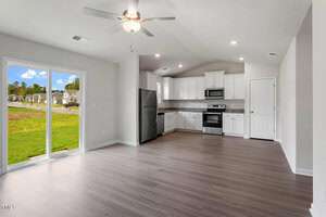 A modern kitchen and living area with light wood flooring. The kitchen features white cabinets, stainless steel appliances, and a granite countertop. A ceiling fan is mounted above. Large sliding glass doors open to a grassy outdoor area with houses visible.