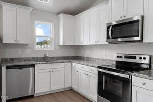 Modern kitchen with white cabinets and granite countertops. Stainless steel appliances include a dishwasher, microwave, and oven. A small window above the sink lets in natural light. Wood flooring contrasts with the white and gray decor.