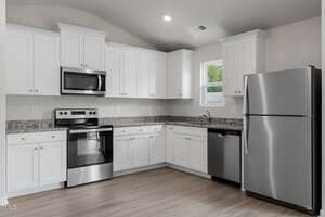 Modern kitchen with white cabinetry, stainless steel appliances including a refrigerator, oven, microwave, and dishwasher. Granite countertops and light wood flooring are present, with a small window above the sink area.