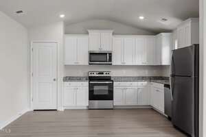A modern kitchen with white cabinets, a microwave, stove, and a refrigerator. The countertop is granite, and the flooring is light wood. The ceiling has recessed lighting, and there is a closed door on the left side.