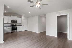 Spacious empty room with light wood flooring and white walls. A ceiling fan is centered in the room. The kitchen area features white cabinets, stainless steel appliances, and granite countertops. An open doorway leads to another room.