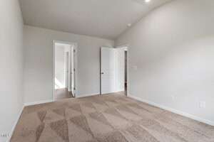 An empty room with light beige walls and carpeted floor. The room features two open doors; one leads to another room with natural light, and the other to a corridor. The ceiling is vaulted and there are no furnishings.