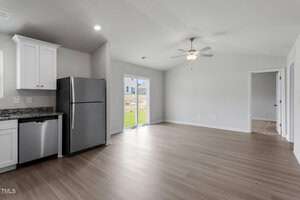 A modern kitchen and living area with light wood flooring. The kitchen has white cabinets, a stainless steel refrigerator, and a dishwasher. A ceiling fan is installed above the living space, and a sliding glass door leads to an outside area.