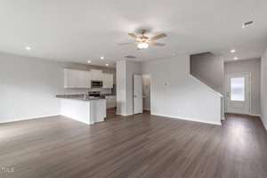 Spacious, empty interior of a modern home featuring an open concept living area. The room has light wood flooring, a ceiling fan, and recessed lighting. The kitchen includes white cabinetry, granite countertops, and stainless steel appliances.