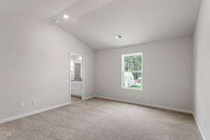 Empty room with beige carpet and light gray walls, featuring a high ceiling and single window. A doorway opens to a glimpse of another room with white cabinetry and natural light.