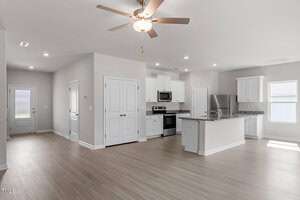 A modern, unfurnished kitchen and living area with light wood flooring. The kitchen features white cabinets, stainless steel appliances, and a granite countertop island. A ceiling fan is installed in the living area, and natural light enters through windows.