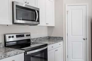 Modern kitchen interior with white cabinets and a granite countertop. Features a stainless steel microwave above an electric stove and oven. A white door with a silver handle is on the right side of the image.