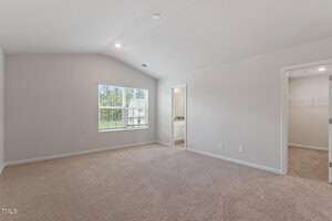Empty room with beige carpet and white walls, featuring a large window allowing natural light, a doorway leading to a bathroom, and an open door to a walk-in closet. Recessed lighting is installed in the ceiling.