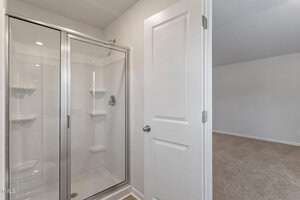 View of a bathroom featuring a glass-enclosed shower with shelves, adjacent to a white door. Beyond the door is a carpeted room with neutral walls.