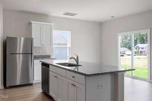 Modern kitchen with white cabinets, a stainless steel refrigerator, and a large island featuring a sink. Light wood flooring and sliding glass doors leading to a lawn are visible. Ceiling lights and a small window offer natural illumination.