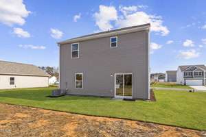 A two-story house with gray siding is shown from the backyard. It has several windows and a glass patio door. The yard is grassy, and an AC unit is visible on the left side. The sky is clear with a few clouds. Other houses are in the background.