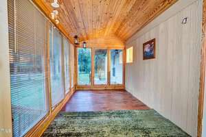 A narrow room with wooden walls and ceiling. One side has large windows with vertical blinds, and the floor is partially carpeted and partially wooden. A doorway leads outside, and a framed picture hangs on the wall.