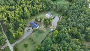 Aerial view of a rural landscape featuring a circular driveway leading to a house surrounded by trees. Adjacent are a small pond and two additional structures. Dense forests border the property on multiple sides.