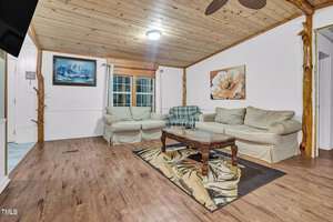 A cozy living room with a wooden ceiling and floor. The space features two beige sofas, a wooden coffee table on a patterned rug, a painting of a flower, and a painting on the wall. A window with curtains is visible in the center.