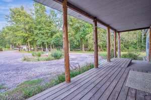 A wooden porch with a view of a gravel driveway and green trees in the background. The porch has wooden beams supporting a roof, and a doormat is placed on the wooden floor. The scene is outdoors, surrounded by nature.