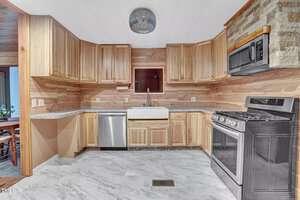 A kitchen with wooden cabinets and a light wood-paneled wall. It features a farmhouse sink, stainless steel appliances including a dishwasher, oven, and microwave, and marble-like tile flooring. There's a textured stone backsplash above the stove.