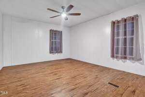 A vacant room with white walls and wooden flooring. It features two windows with sheer brown curtains and a ceiling fan with a light. The room has a simple, clean appearance.