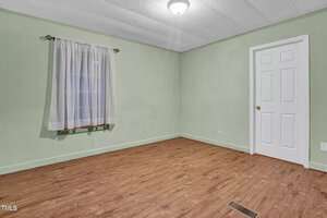 An empty room with pale green walls and wood flooring. A single curtain-draped window is on the left wall. A white closed door is on the right. A ceiling light fixture is centered on the white ceiling. A floor vent is visible on the floor.