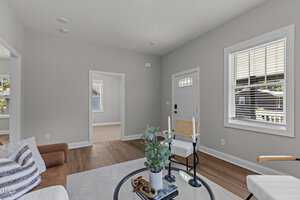 A bright living room with light gray walls and wooden flooring. It features a brown couch with striped cushions, a small round table with plants, a ladder-back chair, and a white front door with windows. A doorway leads to another room.
